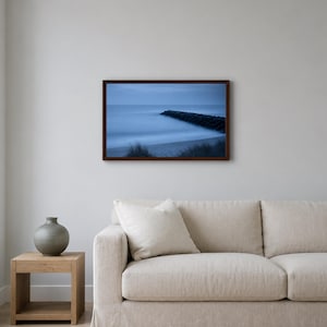 May include: A rectangular wall art print in a dark brown frame featuring a long-exposure landscape photograph of a calm ocean, a sandy beach with dark dune grasses, and a stone jetty extending into the blue-toned water under a twilight sky.