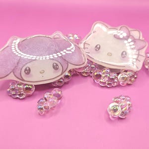 May include: Two character-shaped acrylic charms, one purple and one white, are displayed on a pink surface. The purple charm features a cartoon character with a pearl necklace. The white charm is a cartoon cat with a bow. Iridescent beads surround both charms.