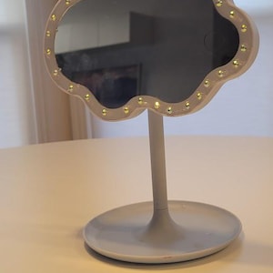 May include: A cloud-shaped vanity mirror with a light gray base and stem. The mirror's frame is a light brown color and is lined with small, golden-yellow lights. The mirror is on a light-colored table.