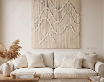 May include: A cream-coloured sofa with cushions, beneath a large macrame wall hanging with a wave pattern. A wooden coffee table and a woven basket are in the room. The overall aesthetic is neutral and cosy, with a focus on natural textures.