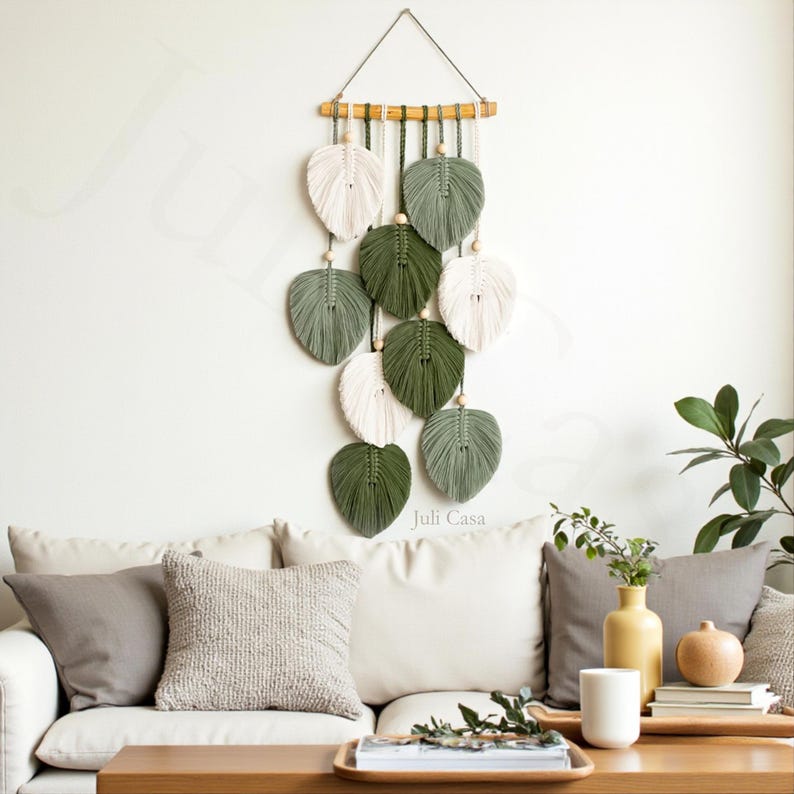 Puede incluir: Colgante de pared de macram&eacute; con dise&ntilde;os en forma de hojas en blanco, verde oliva y verde salvia. Las hojas est&aacute;n suspendidas de un dowel de madera con cuentas de madera. La decoraci&oacute;n de la pared se muestra contra una pared blanca.