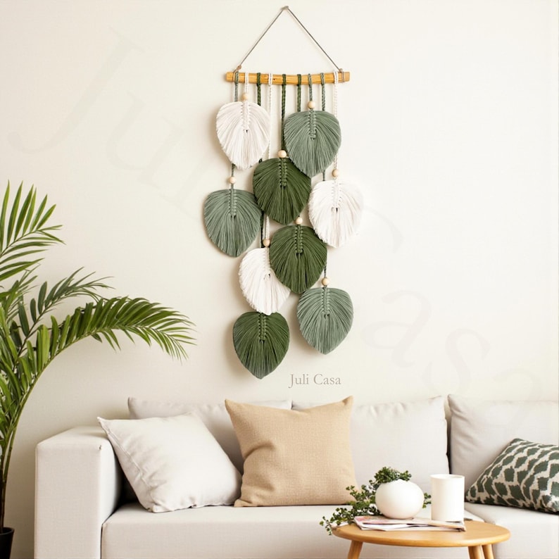 Puede incluir: Un tapiz de pared de macram&eacute; con dise&ntilde;os en forma de hojas en blanco, verde y verde salvia. Las hojas est&aacute;n suspendidas de un pasador de madera con un cord&oacute;n natural. El logotipo de Juli Casa es visible.