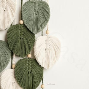 Puede incluir: Colgante de pared de hojas de macram&eacute; en tonos verdes y crema. Las hojas est&aacute;n intrincadamente anudadas y cuentan con cuentas de madera. El logotipo de Juli Casa es visible en la esquina inferior derecha. Decoraci&oacute;n inspirada en la naturaleza.