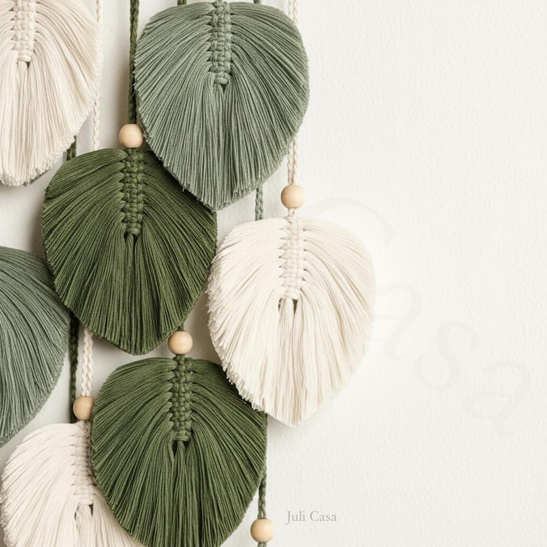 Puede incluir: Colgante de pared de hojas de macram&eacute; en tonos verdes y crema. Las hojas est&aacute;n intrincadamente anudadas y cuentan con cuentas de madera. El logotipo de Juli Casa es visible en la esquina inferior derecha. Decoraci&oacute;n inspirada en la naturaleza.