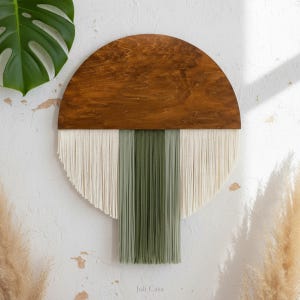 May include: A round wall hanging with a wooden top and a fringe of cream and green yarn. The wooden portion is a half-circle shape with a natural wood grain finish. The yarn fringe is arranged in vertical stripes. The text "Juli Casa" is at the bottom.