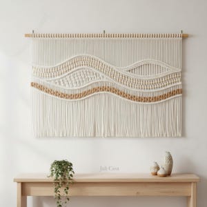 Puede incluir: Tapiz de macramé color crema con diseño de olas. La obra presenta texturas tejidas y anudadas, con una banda horizontal de material tejido en tonos naturales. La pieza está suspendida de un listón de madera, agregando un toque de calidez natural a la decoración.
