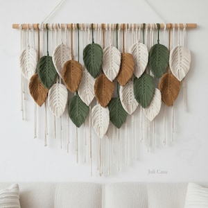 May include: A macramé wall hanging with leaf-shaped designs in white, green, and brown. The leaves are suspended from a wooden dowel with hanging cords, creating a textured, bohemian-style decor piece. The Juli Casa logo is visible.