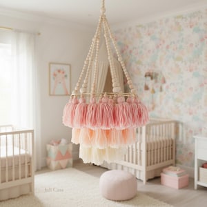 Könnte beinhalten: Eine mehrstufige Hängeleuchte mit rosa, pfirsichfarbenen und cremefarbenen Quasten. Sie hängt an einer Holzperlenstruktur mit einem goldenen Metallring. Das Zimmer hat eine florale Tapete und ein Babybett.