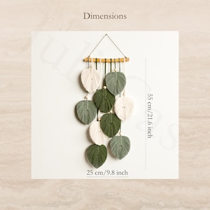 Puede incluir: Colgante de pared de macram&eacute; con dise&ntilde;os en forma de hojas en blanco y verde, suspendido de una clavija de madera. Las dimensiones son 55 cm de altura y 25 cm de ancho. El texto "Dimensions" est&aacute; en la parte superior.