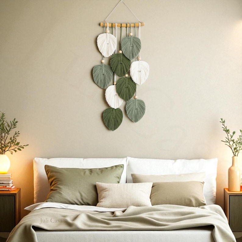 Puede incluir: Un tapiz de pared de macram&eacute; con dise&ntilde;os en forma de hojas en blanco, verde y gris, suspendido de un list&oacute;n de madera. El tapiz se exhibe sobre una cama con almohadas y una manta, creando una est&eacute;tica natural y bohemia.