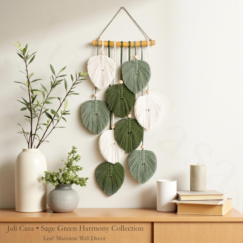 Puede incluir: Un colgante de pared de macram&eacute; con dise&ntilde;os en forma de hojas en blanco, verde salvia y verde oscuro. Las hojas est&aacute;n suspendidas de una clavija de madera con cuentas de madera. El texto "Juli Casa &bull; Sage Green Harmony Collection Leaf Macrame Wall Decor" est&aacute; en la parte inferior.