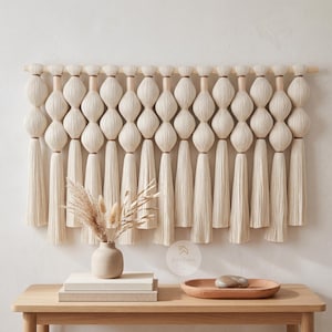 May include: A wall hanging featuring a series of cream-colored yarn spheres suspended from a horizontal wooden rod, with long, straight tassels hanging from the bottom of each sphere. The piece is crafted from off-white fiber and wood.
