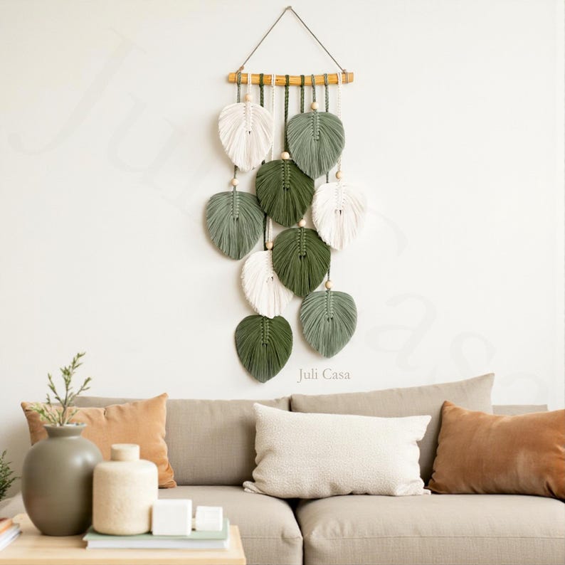 Puede incluir: Un tapiz de pared de macram&eacute; con dise&ntilde;os en forma de hojas en blanco, verde salvia y verde oscuro. Las hojas est&aacute;n suspendidas de una clavija de madera con cuentas de madera. El logotipo de Juli Casa es visible. Decoraci&oacute;n de pared.