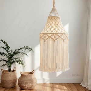 May include: Cream-colored macrame pendant light with a geometric pattern and long fringe. The light hangs from a woven cord and is suspended above a wooden floor. Two woven baskets and a potted plant are in the foreground.
