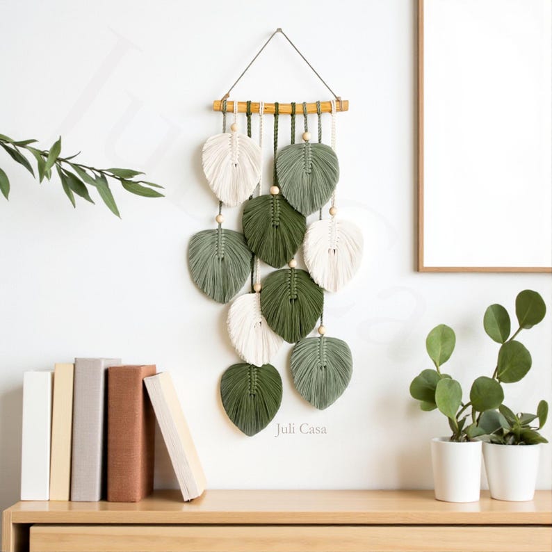 Puede incluir: Un tapiz de pared de macram&eacute; con dise&ntilde;os en forma de hojas en blanco, verde oliva y verde oscuro. Las hojas est&aacute;n suspendidas de una clavija de madera con cuentas de madera. El logotipo de Juli Casa es visible en la pared. El tapiz se exhibe en una pared blanca.