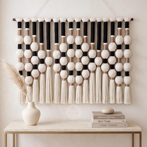 Könnte beinhalten: Ein großer, moderner Wandbehang mit einem sich wiederholenden Muster aus weißen, schwarzen und beigen Elementen. Das Design umfasst große, runde weiße Perlen, schwarze Zylinder und beige Akzente, mit langen weißen Quasten am unteren Ende. Das Stück ist an einer Holzstange aufgehängt.