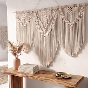 May include: A large, cream-colored macrame wall hanging with a geometric design. The wall decor is suspended from a wooden dowel and features intricate knotting and fringe. A smaller macrame piece is visible in the background. The image also shows a wooden console table with books and a vase.