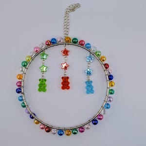 May include: A decorative beaded ornament featuring a silver metal ring with rainbow-coloured beads. Three strands of star and gummy bear charms hang from the ring. A chain is attached for hanging, creating a vibrant, playful design.
