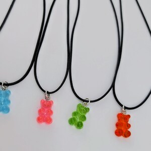 Peut inclure: Quatre colliers avec des pendentifs en forme d'oursons en gélatine. Chaque collier a un cordon noir et un fermoir argenté. Les pendentifs sont bleus, roses, verts et oranges. Les colliers sont présentés sur fond blanc.