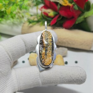 May include: A silver pendant with an oval-shaped, multi-colored stone. The stone features a mix of brown, yellow, and black hues. The pendant has a decorative silver frame with a loop for a chain. The text "Elevate Art Creations" is visible.