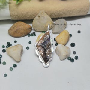 May include: A silver pendant with a large, irregularly shaped, polished stone. The stone has a mix of brown, purple, and white colors. The pendant is surrounded by small, decorative rocks and beads. The text "Elevate Art Creation" is visible.