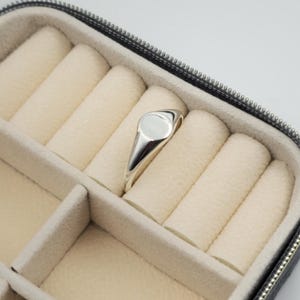 Op de afbeelding: Een gepolijste zilveren zegelring met een glad, rond oppervlak en een taps toelopende band. De ring is geplaatst in een sieradendoosje met beige, met fluweel beklede ringrollen die zijn ontworpen om kleine accessoires veilig te bewaren.