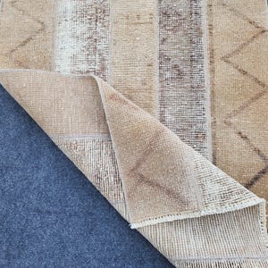 Pode incluir: Um tapete retangular com um canto dobrado, apresentando um design de patchwork em tons de bege, castanho e creme. O tapete tem listras verticais e um padrão em zigue-zague em um lado. O tapete é feito de um material tecido.