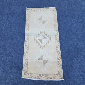 May include: A rectangular area rug featuring a light beige base color with a repeating geometric pattern in a darker tan shade. The design includes a central diamond motif and a decorative border, finished with short white fringe on both ends.