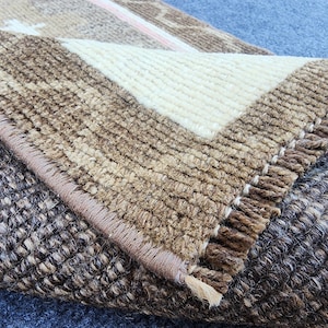 May include: A woven area rug featuring a folded corner that reveals a thick, textured pile. The rug displays a color palette of deep brown, tan, and cream, with a visible stitched edge and a dense, looped construction throughout the surface.