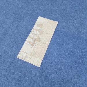 Pode incluir: Um tapete retangular bege e castanho com padrões geométricos. O tapete é colocado sobre uma superfície azul. O design inclui formas e padrões repetidos, criando uma estética vintage ou tradicional.