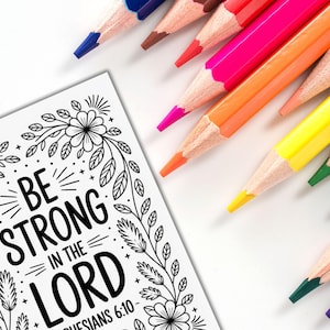 May include: A coloring page with the text "BE STRONG IN THE LORD" and floral border is next to a collection of sharpened colored pencils in various colors. The pencils are arranged diagonally across a white surface, ready for use.