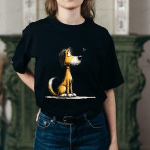 May include: Black t-shirt featuring a cartoon dog with brown and yellow fur, sitting and looking at a small insect. The dog has a white tail and paws, and a red collar. The graphic is centered on the shirt.