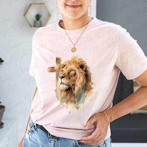 May include: A light pink short-sleeve t-shirt featuring a central graphic of a lion's head with a cross silhouette behind it. The lion is rendered in shades of brown and gold with watercolour-style accents in blue and green tones.