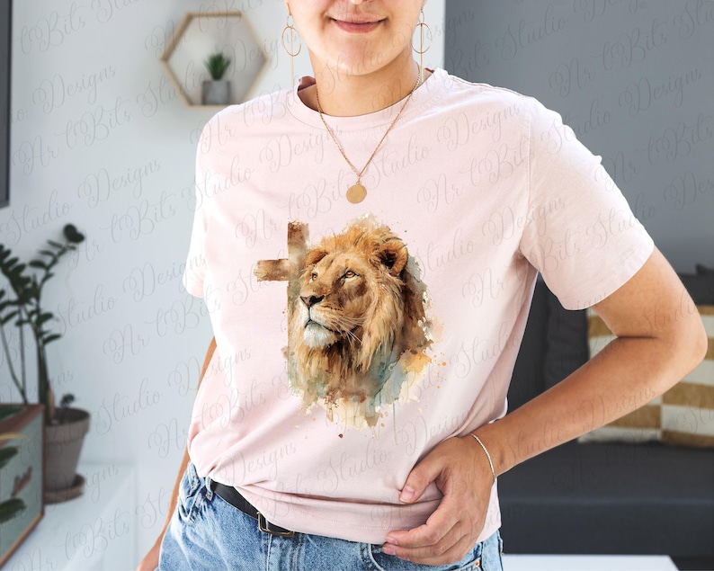 May include: A light pink short-sleeve t-shirt featuring a central graphic of a lion's head with a cross silhouette behind it. The lion is rendered in shades of brown and gold with watercolour-style accents in blue and green tones.