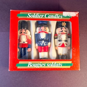 May include: A red rectangular box containing three soldier-shaped candles. The box features the text Soldier Candles and Bougies soldats. The candles are painted in red, blue, white, and black, each wearing a tall hat and a uniform with detailed trim.