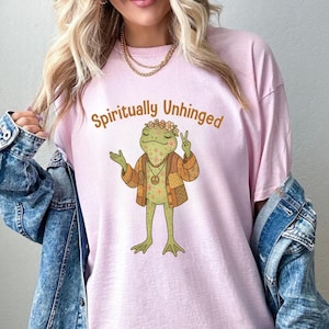 May include: A light pink t-shirt with the words "Spiritually Unhinged" above a cartoon frog wearing a floral crown, jacket, and peace sign. The frog is green and tan. The shirt is worn with a denim jacket.
