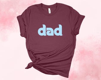 May include: Burgundy t-shirt with the word "dad" in light blue, stylized lettering. The shirt is knotted at the bottom left. The background is a watercolor wash of pink and white.