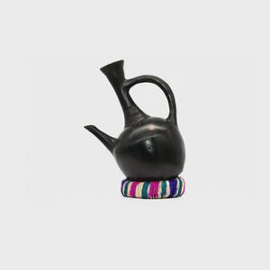 Könnte beinhalten: Eine schwarze Keramik-Kaffeekanne mit gebogenem Griff und Ausguss, die auf einem farbenfrohen gewebten Sockel steht. Die Kanne hat einen bauchigen Körper und einen langen, schlanken Hals. Der Sockel hat Streifen in Rosa, Blau, Grün und Weiß.