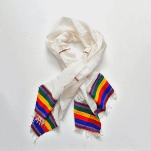 Può includere: Una sciarpa bianca con estremità a righe colorate. La sciarpa ha un design semplice con una trama strutturata. Le estremità presentano vivaci bande di strisce rosse, gialle, verdi e blu, aggiungendo un tocco di colore.