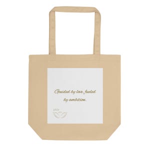 May include: Beige tote bag with a rectangular white panel. The panel features the text "Guided by love, fueled by ambition" in gold script, along with a small gold illustration of a bird. The bag has a simple design with a rectangular shape and handles.