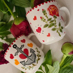 Könnte beinhalten: Zwei weiße Keramikbecher mit handgemalten Designs. Ein Becher zeigt eine rote Rose und grüne Blätter, der andere einen Anker und ein Herz. Beide haben rote Herzakzente und einen gewellten Rand. Die Becher sind von roten Pfingstrosen und grünen Blättern umgeben.