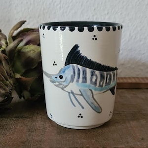 Könnte beinhalten: Weißer Keramikbecher mit handgemaltem blauem Marlin-Design. Der Becher hat einen schwarzen Rand und kleine schwarze Punkte an der Oberseite und den Seiten. Im Hintergrund befindet sich eine Artischocke, auf einer Holzoberfläche.