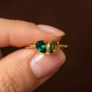 May include: A gold ring featuring two pear-cut gemstones set side by side to form a heart shape. One gemstone is a deep emerald green, while the other is a lighter olive green. The band is thin and polished gold, creating a delicate appearance.
