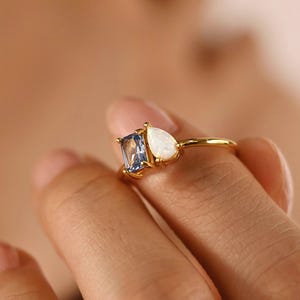 May include: A gold ring featuring a rectangular blue gemstone and a pear-shaped white gemstone. The ring is held by a hand, showcasing the delicate design and the contrast between the gemstones and the gold band. The ring is a piece of jewelry.