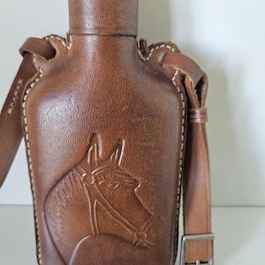 Könnte beinhalten: Braune Lederflasche mit einem Pferdekopf-Design. Die Flasche hat einen Schulterriemen und eine Metallschnalle. Die Kanten sind mit weißem Faden genäht. Die Oberseite der Flasche hat einen dekorativen Rand.