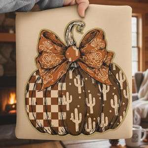 May include: A decorative pillow featuring a brown pumpkin design with a large bow. The pumpkin has a checkerboard pattern and cactus illustrations. The bow is a paisley design in shades of brown and orange. The pillow is held by a person.