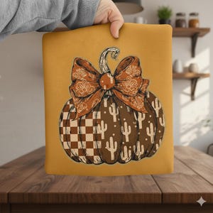 May include: A decorative pillow with a pumpkin design. The pumpkin is brown with a checkered pattern and cactus illustrations. It features a large orange bow and a curved stem. The pillow is held by a person.