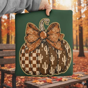 May include: A green pillow with a brown pumpkin design. The pumpkin has a large bow with a paisley pattern, a checkered pattern, and cactus illustrations. The pillow is held by a person.