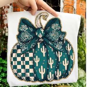 May include: A white t-shirt featuring a large graphic of a teal pumpkin with a gold glitter outline. The pumpkin has a checkered pattern on one side, a cactus print on the other, and a large teal bow with floral details at the top stem.