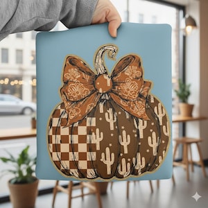 May include: A decorative pillow featuring a brown pumpkin design with a large bow. The pumpkin has a checkered pattern and cactus illustrations. The background is a light blue color.